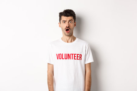 Shocked and confused man in volunteer t-shirt staring at camera speechless, standing against white backgroundの写真素材