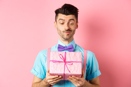 Holidays and celebration concept. Happy smiling guy receive white day gift from his valentine, looking at present with intrigued face, standing over pink backgroundの写真素材