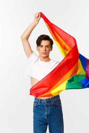 Vertical view of happy queer person in crop top and jeans waving raised rainbow flag, celebrating lgbtq holiday, standing over white backgroundの写真素材
