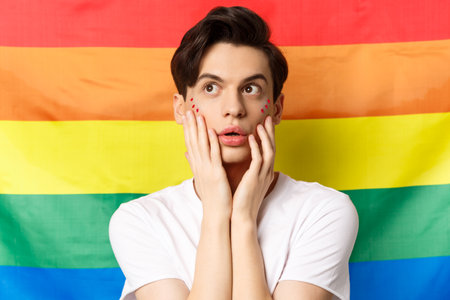 Portrait of sensual and beautiful gay man with glitter makeup on face, looking at upper left corner excited, standing against lgbtq rainbow flagの写真素材
