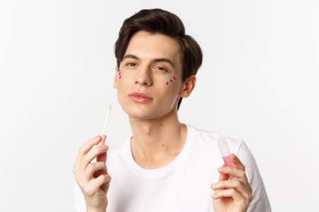 Close-up of beautiful queer person with glitter on face applying lips gloss and looking self-assured at camera, standing over white backgroundの写真素材