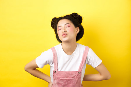 Romantic korean girl close eyes and pucker lips, waiting for sweet kiss on Valentines day, kissing lover, standing on yellow backgroundの写真素材