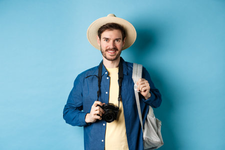 Cheerful handsome guy going on vacation, wearing summer hat and holding backpack with camera for photos, smiling excited of holiday, standing on blue backgroundの写真素材