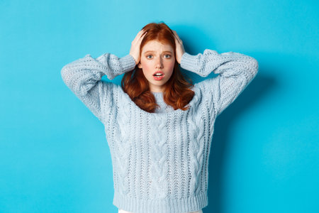 Nervous redhead girl standing overwhelmed, holding hands on head in panic and staring at camera, standing anxious against blue backgroundの写真素材
