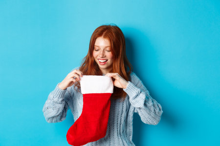 Winter holidays and gifts concept. Funny redhead girl looking inside Christmas stocking and smiling happy, receiving xmas present, standing against blue backgroundの写真素材