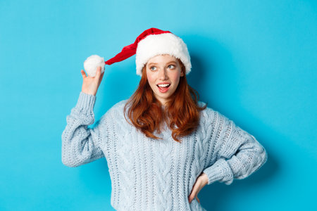 Winter holidays and Christmas Eve concept. Silly redhead girl with freckles, touching her santa hat and thinking, planning New Year celebration, standing over blue backgroundの写真素材