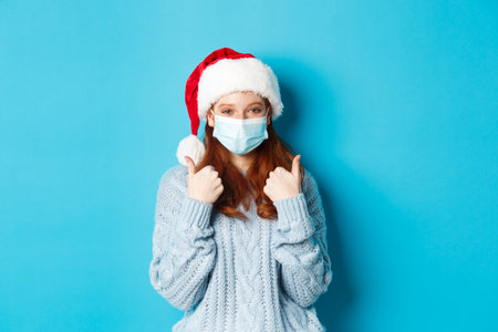 Christmas, quarantine and covid-19 concept. Cute teen redhead girl in santa hat and sweater, wearing face mask from coronavirus, showing thumbs up, standing over blue backgroundの写真素材