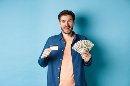 Smiling caucasian guy showing plastic credit card and money, looking happy, standing on blue...