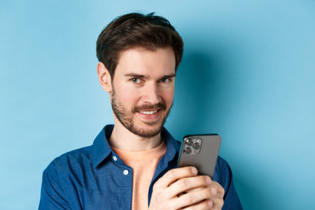 Close-up of attractive caucasian man holding mobile phone and smiling at camera, standing in casual outfit on blue backgroundの写真素材