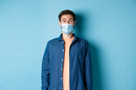 Covid-19 and healthcare concept. Image of handsome modern guy in face mask raising eyebrows in surprise, looking at something in awe, blue backgroundの写真素材