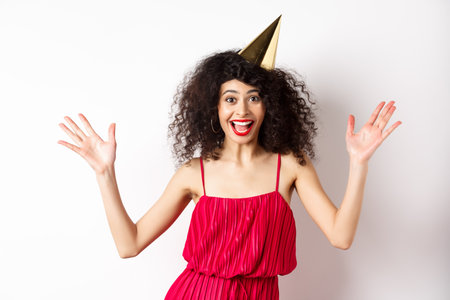 Cheerful young woman in red dress, celebrating birthday, wearing party hat and smiling, screaming of joy, standing on white backgroundの写真素材