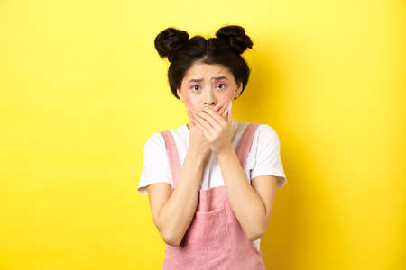 Worried asian teen girl covering mouth with hands, looking concerned and anxious, standing scared on yellow background with glam makeup and summer clothesの写真素材