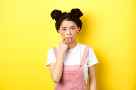 Rude asian girl stretch eyelid and showing tongue, mocking someone, standing on yellow backgroundの写真素材