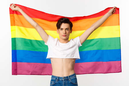 Out and proud. Happy and confident gay man with glitter on face raising lgbtq rainbow flag and smiling at camera, standing in crop top, white backgroundの写真素材