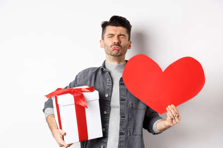Sad and heartbroken man being rejected, crying and holding red heart with gift box, breakup on Valentines day, white background. Love and relationship conceptの写真素材