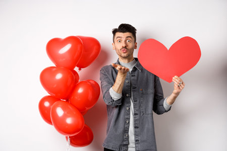 Valentines day and love concept. Romantic boy showing heart card and blowing air kiss at camera, looking with sympathy at lover, celebrating white dayの写真素材