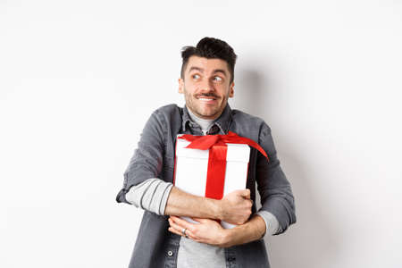 Happy boyfriend hugging gift from lover, looking aside at empty space and smiling excited, making romantic present on valentines day, standing on white backgroundの写真素材