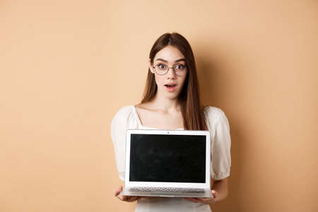 Surprised young woman in glasses showing laptop screen with amazed face, standing on beige backgroundの写真素材