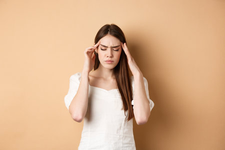 Young woman having headache, touching head temples with closed eyes and tensed face, standing with painful migraine on beige backgroundの写真素材