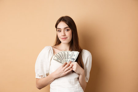 Smiling rich woman showing dollar bills and looking happy at camera, making money or fast loan cash, standing on beige backgroundの写真素材