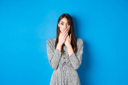 Shocked woman in dress gasping, covering mouth with hands and look excited, hear gossips, standing against blue backgroundの写真素材