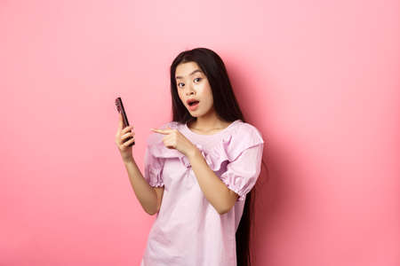 Excited beautiful asian woman, pointing finger at smartphone and look curious, checking out online promo, standing against pink backgroundの写真素材