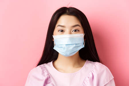 Covid-19 and healthy people concept. Close-up of asian cute girl wearing medical mask during virus outbreak, standing against pink backgroundの写真素材