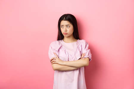 Sad and gloomy asian woman look away , sulking from unfair situation, cross arms on chest disappointed, standing against pink backgroundの写真素材