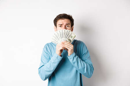 Worried young man looking with pleading face, showing money, standing in blue sweatshirt over white backgroundの写真素材