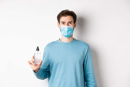 health and quarantine concept. Young caucasian man advice hand sanitizer, showing antiseptic and wearing medical mask, standing on white backgroundの写真素材