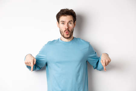 Amazed young man with beard, standing in casual blue shirt, saying wow and pointing down, look here gesture, standing over white backgroundの写真素材