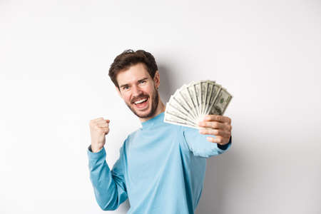 Happy caucasian man stretch out hand with money in dollars, saying yes and celebrating income, got cash prize, standing over white backgroundの写真素材