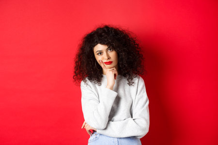 Bored and tired curly woman with red lips, looking bothered at camera, standing against studio...
