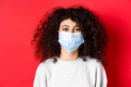 Covid-19 and pandemic concept. Close-up of modern young woman with curly hair, wearing medical mask from coronavirus, smiling at camera, red backgroundの写真素材