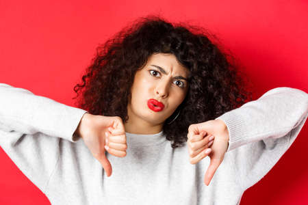 Close-up of disappointed young woman with curly hairstyle, showing thumbs down and frowning, condemn something bad, standing displeased on red backgroundの写真素材