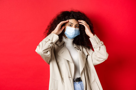 Pandemic and quarantine concept. Stylish young woman with curly hair, going outside in medical mask...