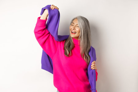 Carefree korean elderly woman in pink sweater, dancing with sweatshirt on shoulders and smiling, posing happy over white backgroundの写真素材