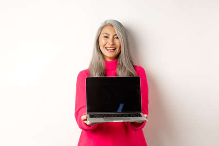 E-commerce concept. Beautiful asian senior businesswoman showing blank laptop screen and smiling, standing in pink sweater over white backgroundの写真素材