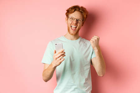 Happy redhead guy in glasses and t-shirt winning online prize, shouting yes with joy and satisfaction, holding smartphone and making fist pump, pink backgroundの写真素材