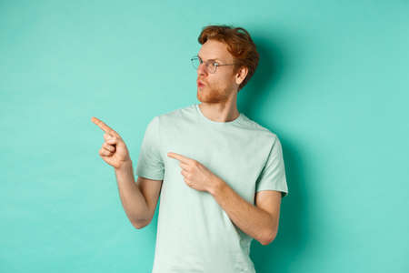 Impressed redhead man in glasses and t-shirt, pointing fingers and looking left at promo offer, staring happy, standing over turquoise backgroundの写真素材