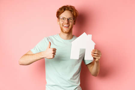 Real estate. Cheerful man in glasses and t-shirt recommending broker agency, showing paper house cutout and thumbs-up, standing over pink backgroundの写真素材