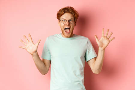 Angry and pressured young man losing temper, spread hands sideways and screaming with furious face, standing in glasses and t-shirt against pink backgroundの写真素材