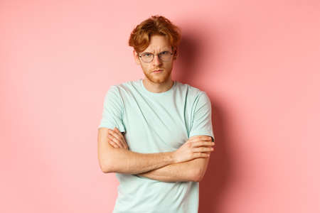 Angry man in glasses with red hair frowning, cross arms on chest in defensive pose, sulking at you, standing over pink backgroundの写真素材