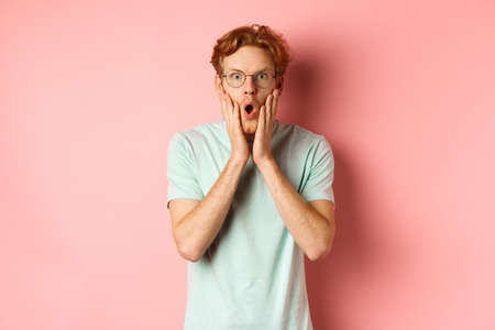 Shocked redhead man gossiping, staring impressed at camera and touching face, standing in glasses against pink backgroundの写真素材