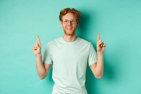 Image of handsome bearded man with red hair, wearing t-shirt and glasses, smiling happy and pointing fingers up, showing promo offer, standing over turquoise backgroundの写真素材