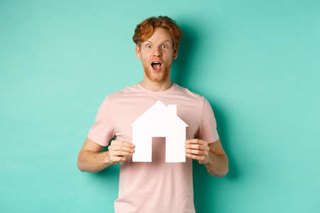 Real estate concept. Excited redhead guy with beard, showing paper house cutout and gasping in awe, staring fascinated at camera, standing over mint backgroundの写真素材