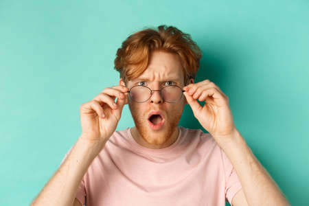 Close up portrait of redhead guy take-off glasses and gasping startled, looking shocked at camera, standing over turquoise backgroundの写真素材
