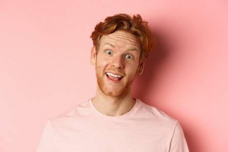 Headshot of funny redhead guy showing tongue, making silly faces at camera, standing joyful against pink backgroundの写真素材
