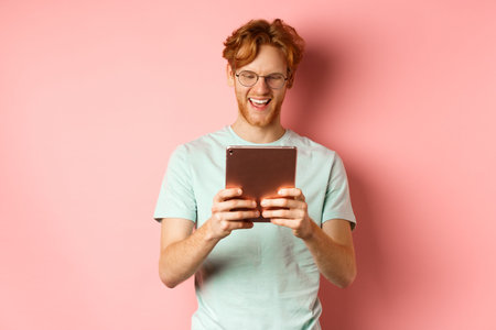 Happy guy with red hair and beard using digital tablet, reading screen and smiling, standing over...