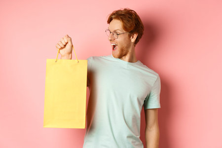 Handsome young man buying presents, holding shopping bag and looking amused, standing over pink...